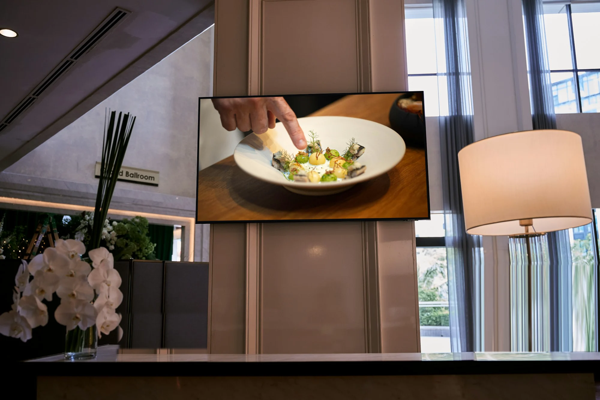 A hotel lobby with orchids and a lamp, its TV on a cooking show