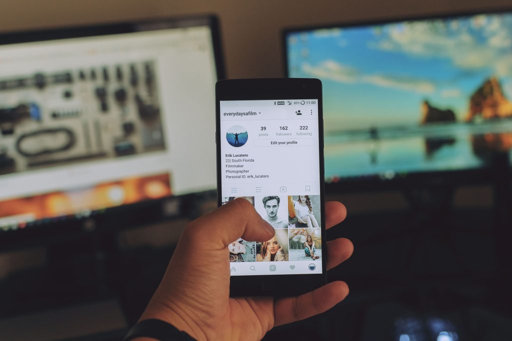 A hand holding a phone showing a photo feed, with a laptop behind it