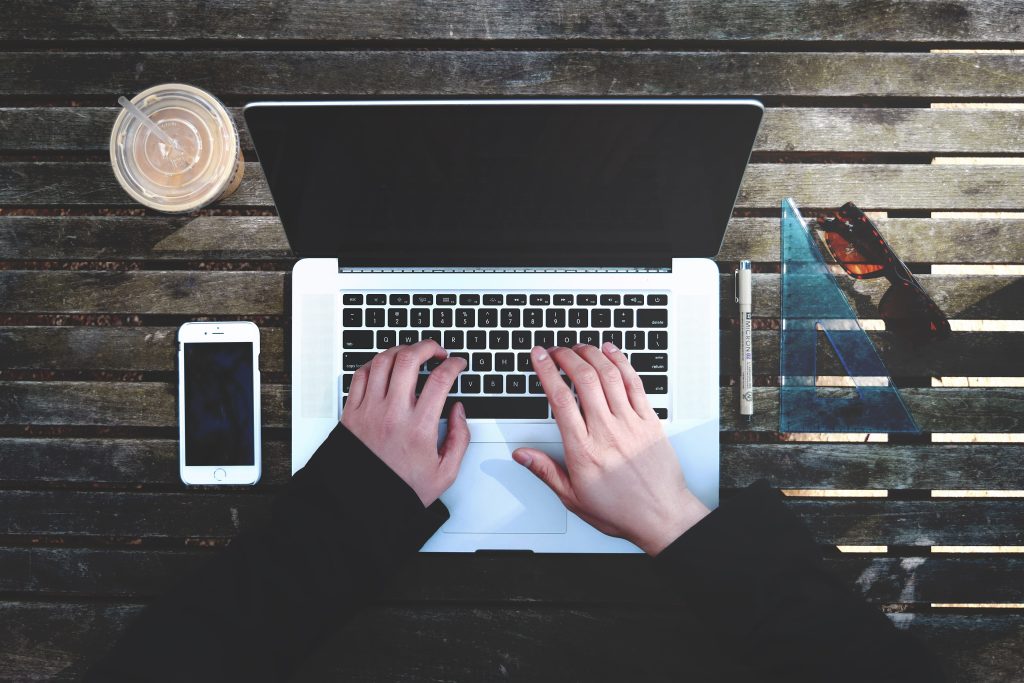 Hands typing on a laptop beside a phone and a notebook on a wooden desk