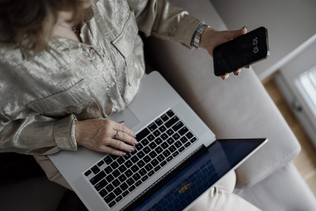 A person on a sofa with a laptop on their lap, holding a smartphone in one hand
